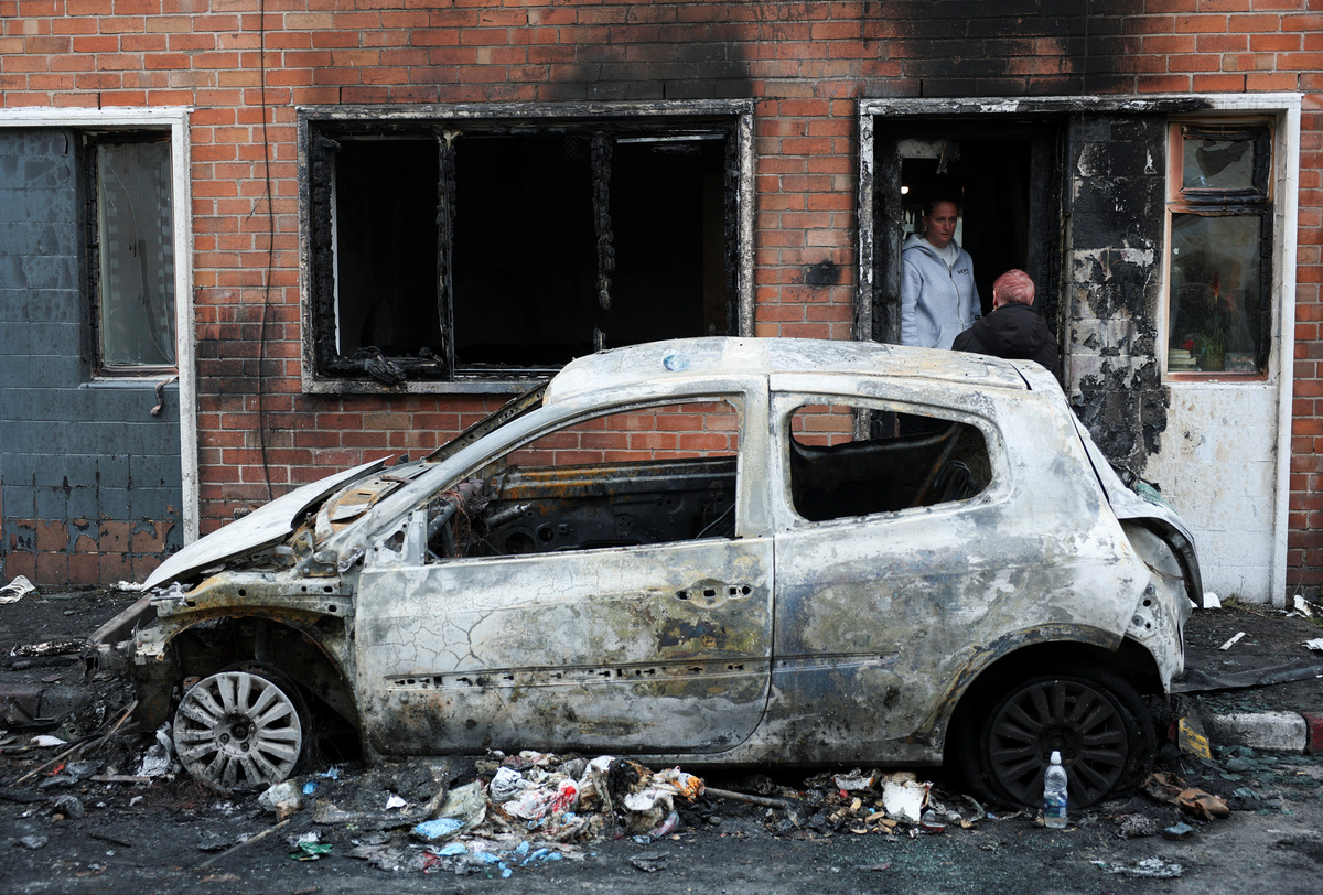 A woman speaks to a person from the doorway of her burnt house, as a destroyed car stands on the street, in the aftermath of anti-immigrant protests following a knife attack on June 8, which left a man seriously injured and prompted police to declare a critical incident, in Belfast, Northern Ireland, June 10, 2026. REUTERS/Isabel Infantes