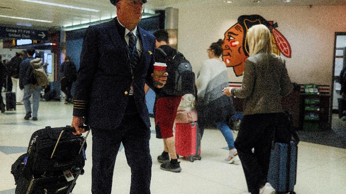 A pilot carries a suitcase and a coffee cup while walking past travelers near a Chicago Blackhawks logo as flight delays persist amid FAA measures more than a month into the ongoing U.S. government shutdown, at O’Hare International Airport in Chicago, Illinois, U.S., November 10, 2025. REUTERS/Chris Helgren     TPX IMAGES OF THE DAY