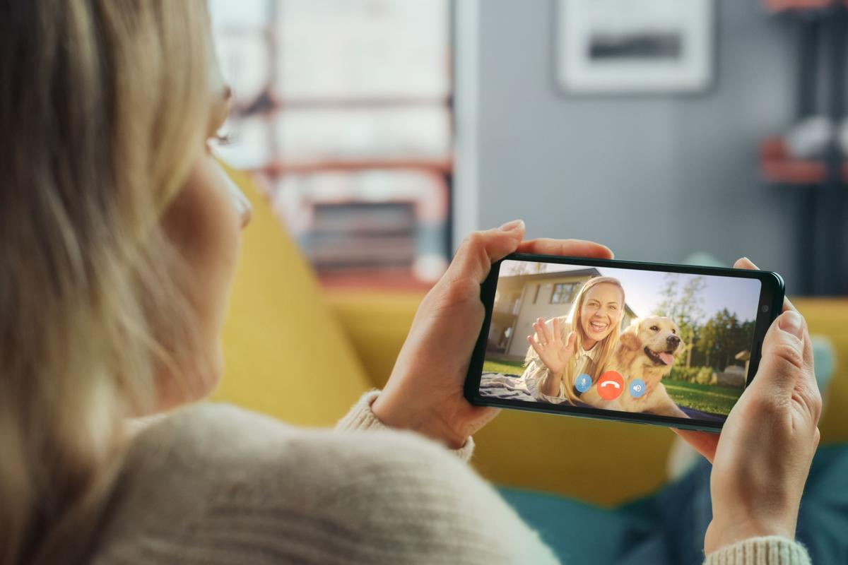  A stock image of a woman holding her phone and video calling with another person and a dog. 