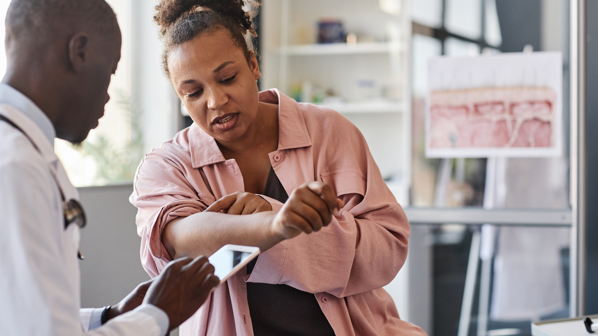 A stock image of a woman suffering from psoriasis visiting dermatologist in clinic and pointing on rash spots on arms.