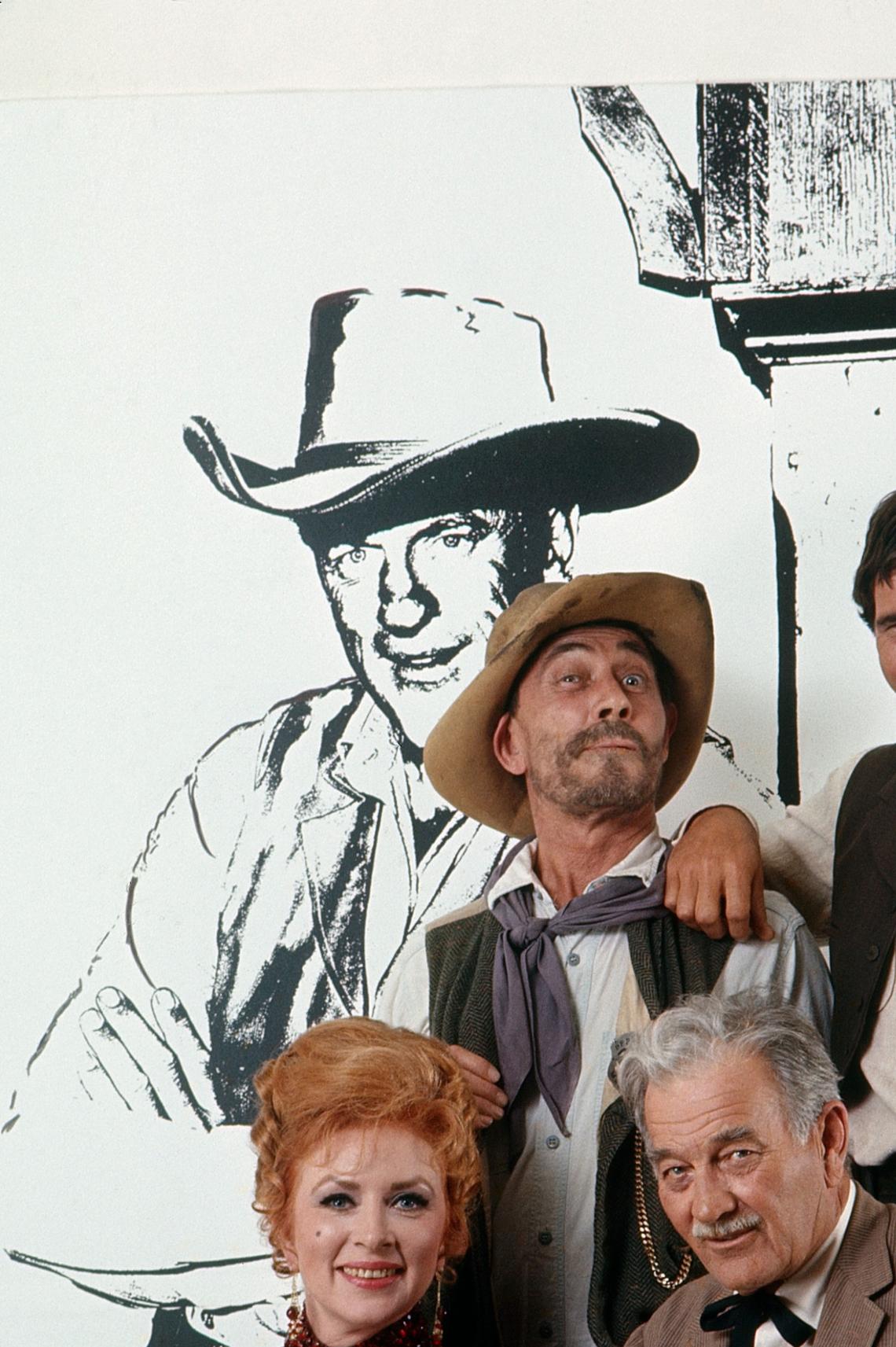 GUNSMOKE, from left: Amanda Blake, Ken Curtis, Milburn Stone, Buck Taylor in front of an illustration of James Arness (1970)Gene Trindl/TV Guide/courtesy Everett Collection
