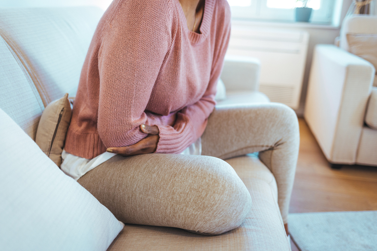  Stock image: a young woman sits down on her couch, suffering from period pains at home. 