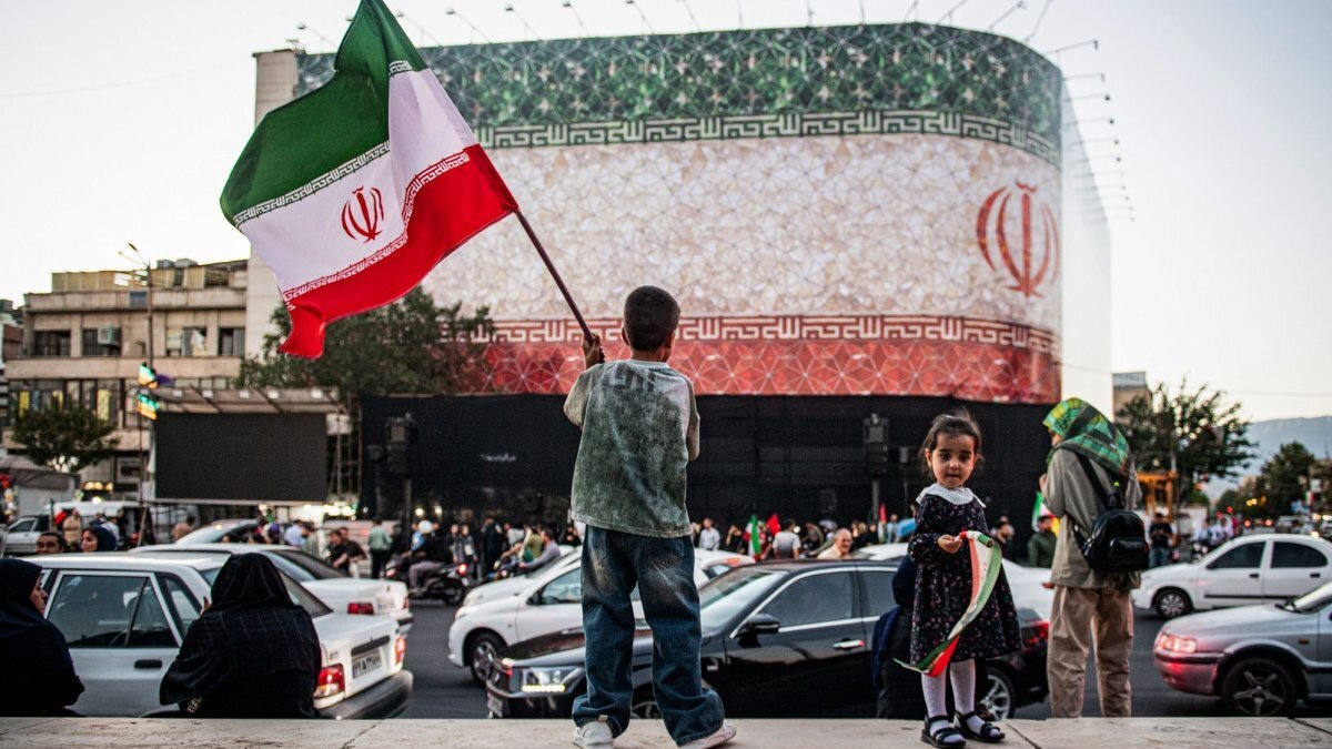 An Iranian child carries the national flag as the Iranian flag waves atop a building on Sunday. President Donald Trump and Pakistani Prime Minister Shehbaz Shafir announced Sunday that the United States and Iran have come to a peace agreement. Photo by Behnam Tofighi/UPI