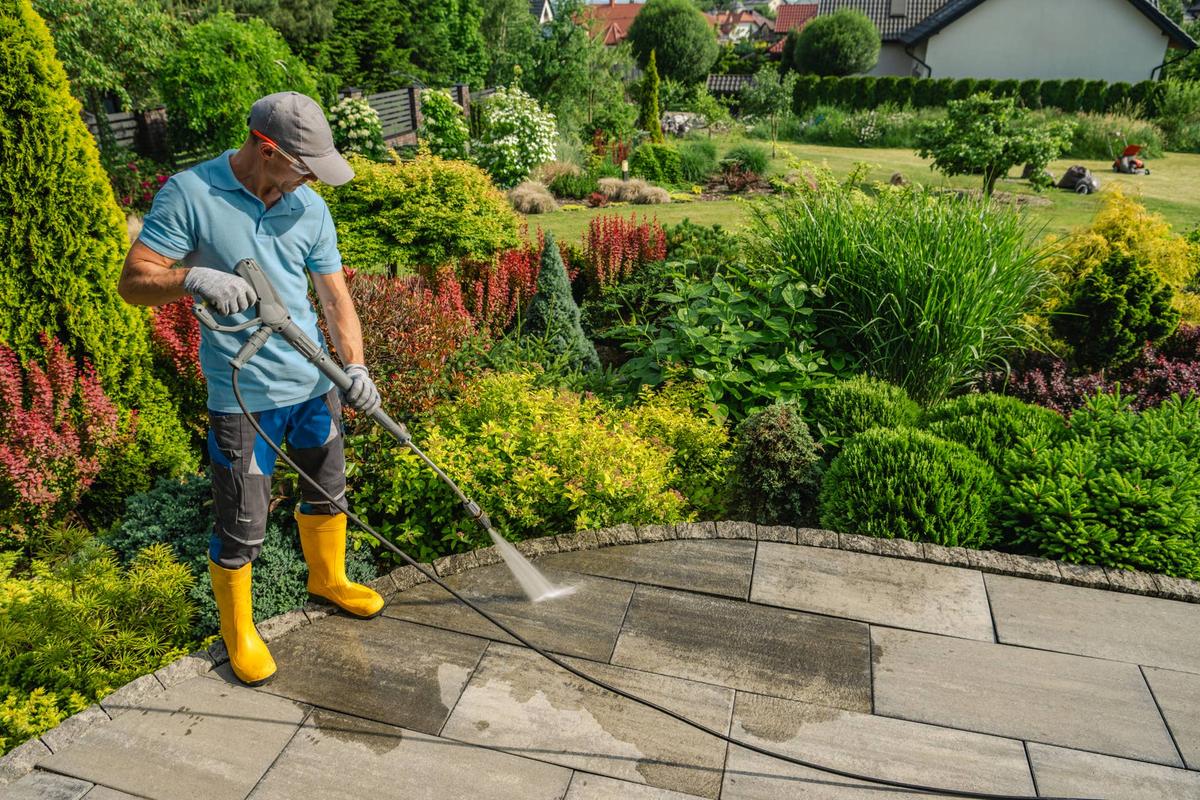  A man in yellow boots uses a pressure washer on a stone patio in a vibrant garden full of plants and flowers.