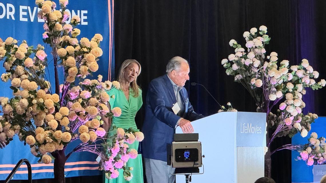 LifeMoves Board Chair Melissa Selcher stands behind John A. Sobrato as he speaks at "A Home For Everyone," a benefit luncheon for LifeMoves held at the Crowne Plaza Palo Alto on Thursday, April 23, 2026. (Sal Pizarro/Bay Area News Group)