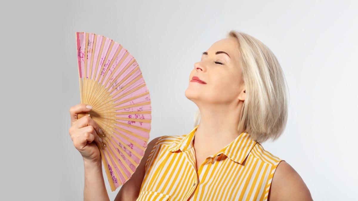 Attractive middle aged woman relaxing with fan. Mature woman experiencing hot flush from menopause isolated on grey.