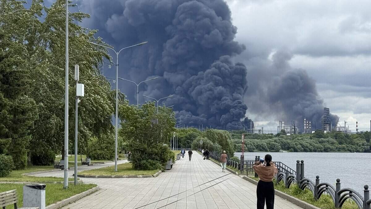People walk in a park as black smoke rises from the area of the Russian oil producer Gazprom Neft's oil refinery on the south-eastern outskirts of Moscow on June 18, 2026.