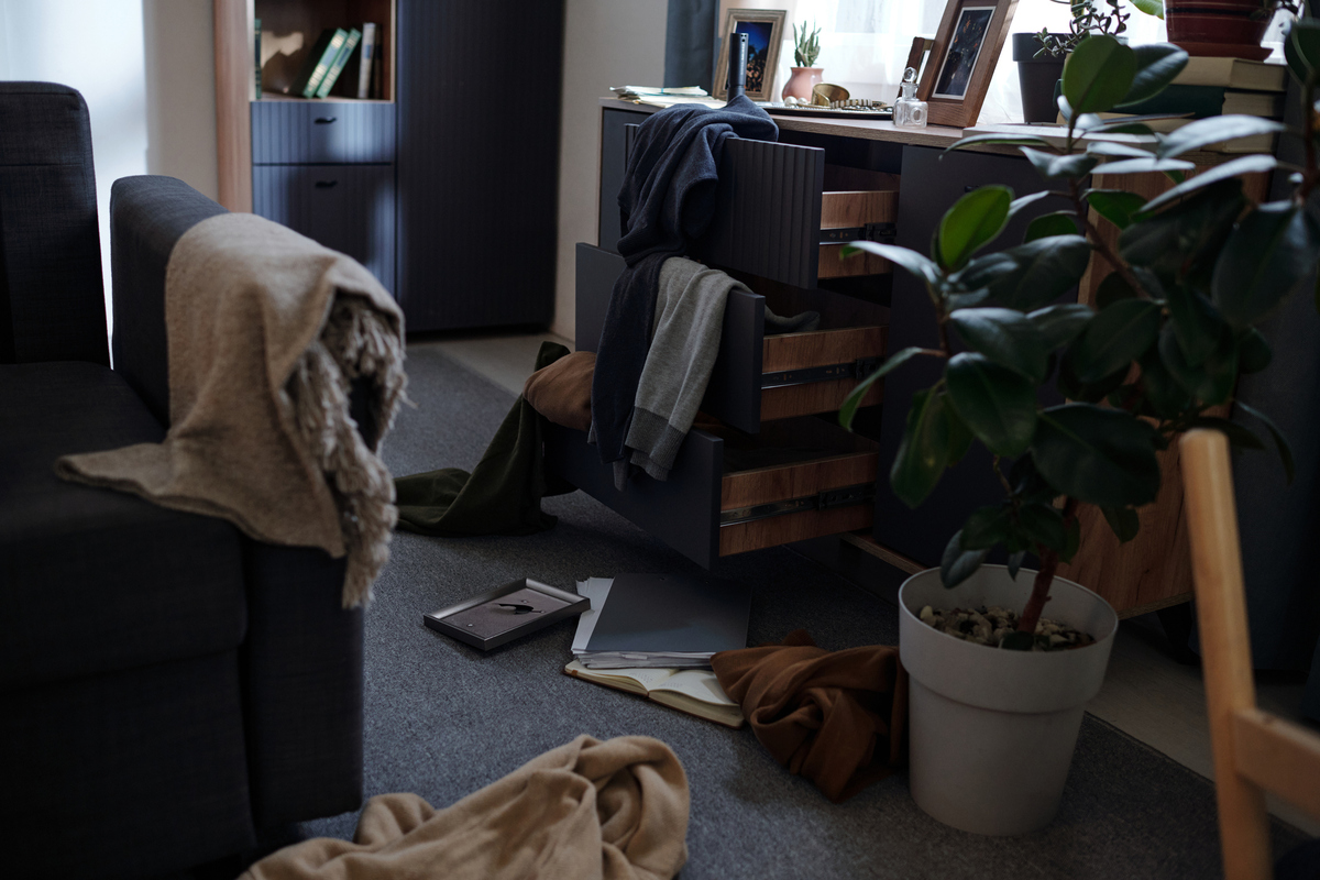  A stock image of a messy living room scene showing open drawers with scattered clothing and papers on floor, potted plant in foreground. 