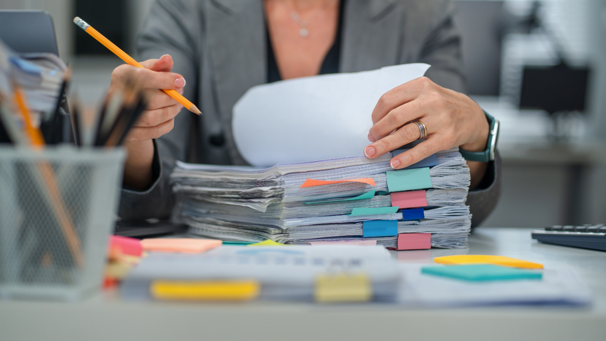 Senior businesswoman, searching documents in messy stack. Reports, forms, file folders, ordering and organization. Frustration, emotional stress, business office overflow. 