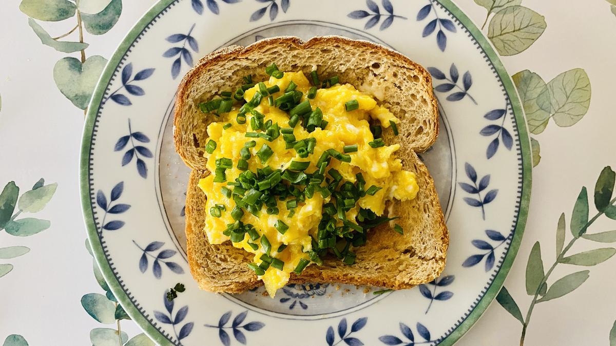 High angle closeup view of golden scrambled organic eggs with fresh chives on a slice of buttered, toasted sourdough bread on a round, leaf pattern ceramic plate