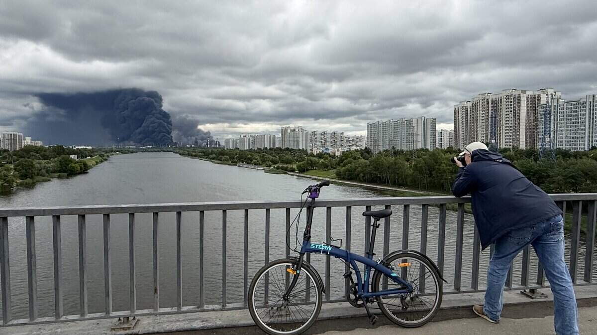 A man takes photographs from a bridge across the Moskva river as black smoke rises from the area of the Russian oil producer Gazprom Neft's Moscow oil refinery on the south-eastern outskirts of Moscow on June 18, 2026.