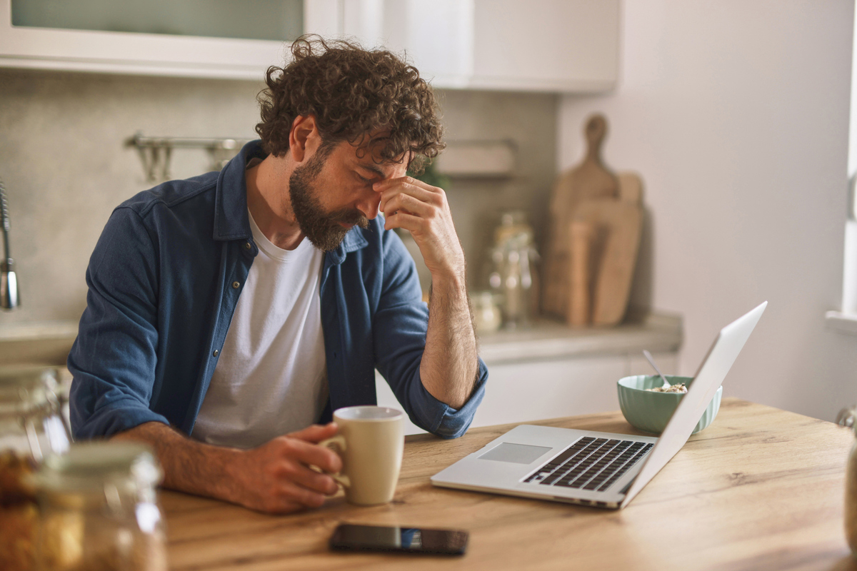  Stock image: a man sits at a table with a laptop and mug, looking stressed. 