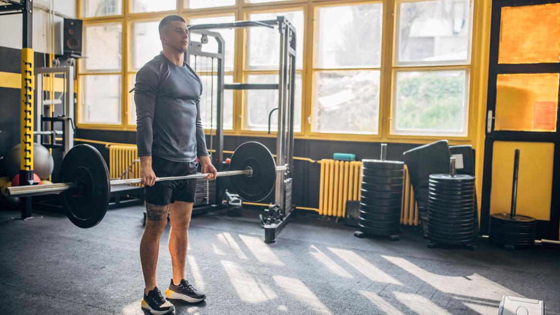 A man wearing a gray long-sleeve shirt and shorts is lifting weights in a gym with large windows, surrounded by exercise equipment and weights..