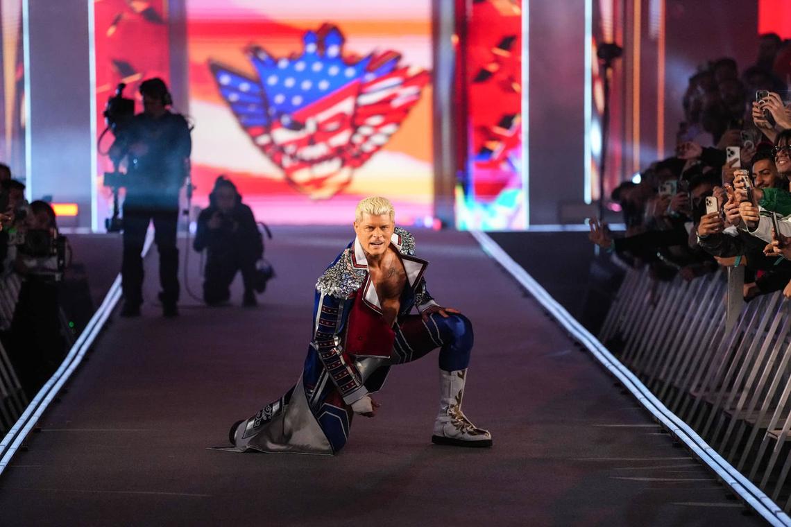  Cody Rhodes will defend the Undisputed WWE Championship against Randy Orton in a dream match turned sour with Pat McAfee's involvement. Professional wrestler Cody Rhodes performs his signature entrance on a long, dark ramp leading to a ring. He is kneeling on one knee, looking forward with an intense expression. He wears an elaborate blue, red, and silver "American Nightmare" themed trench coat with silver sequins on the shoulders, paired with white and gold wrestling boots. In the background, a large screen displays his logo—a skull with the American flag—while fans line the barricades holding up phones to take photos.