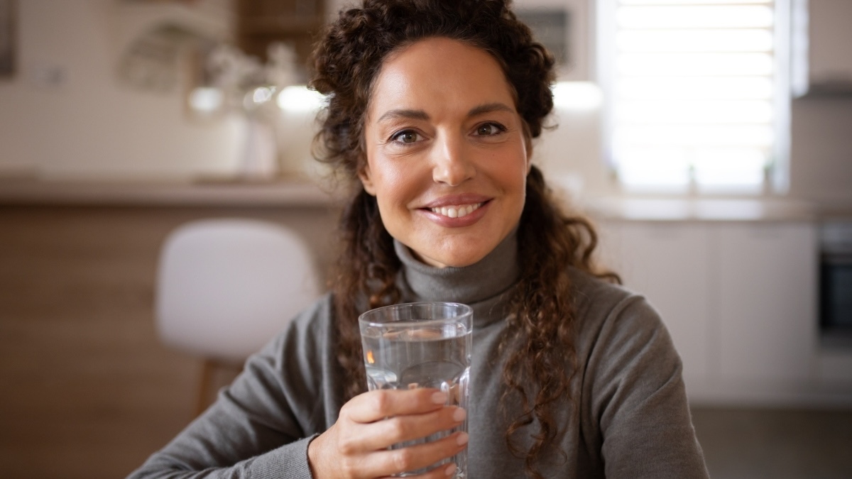 A cheerful woman enjoys a moment of wellness, holding a glass of water with a bright smile, perfect for health campaigns, hydration tips, or morning routine content.