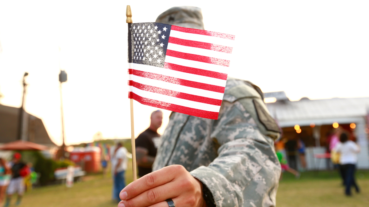 U.S. veteran holding American flag. Anonymous army solider in National Guard wearing military uniform. Freedom concept for armed forces, military pride, patriotism on independence day / July 4th / Fourth of July or Veteran's Day. Waist up candid portrait or real life real people outdoors during summertime at a county fair in America.