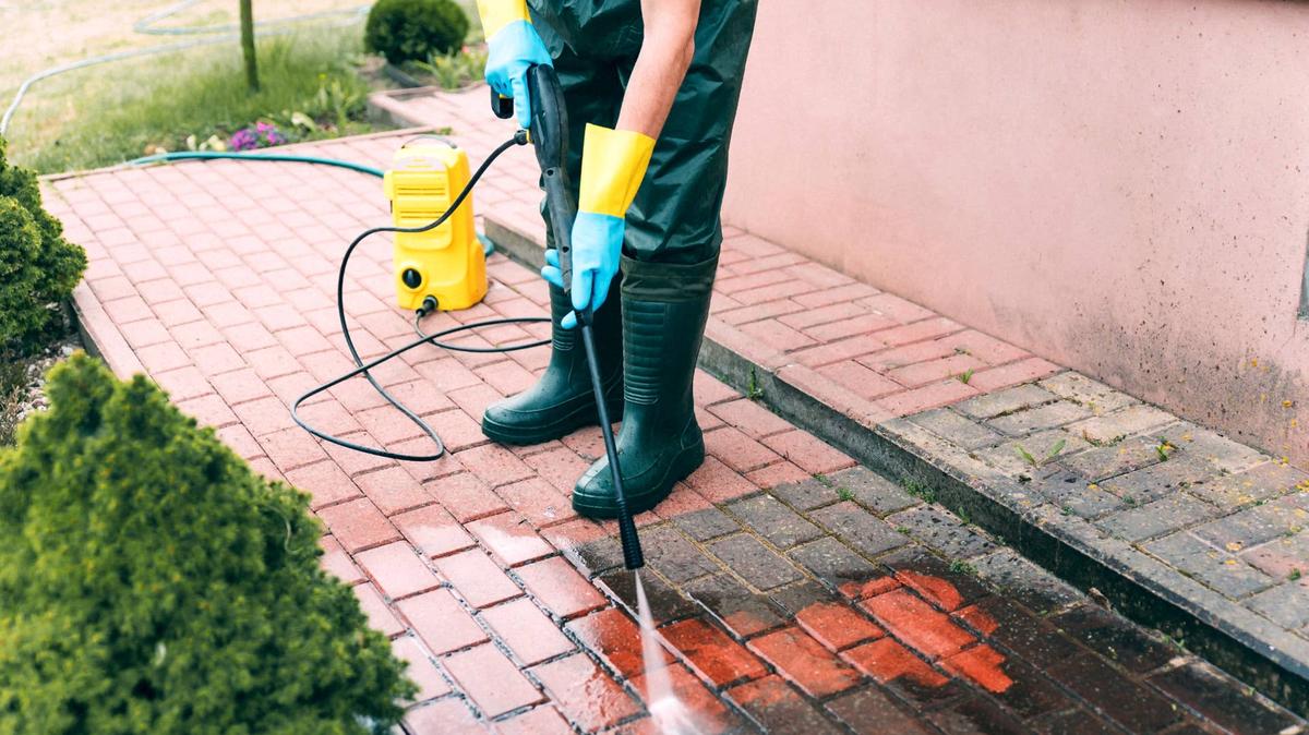 A man in yellow boots uses a pressure washer on a stone patio in a vibrant garden full of plants and flowers.