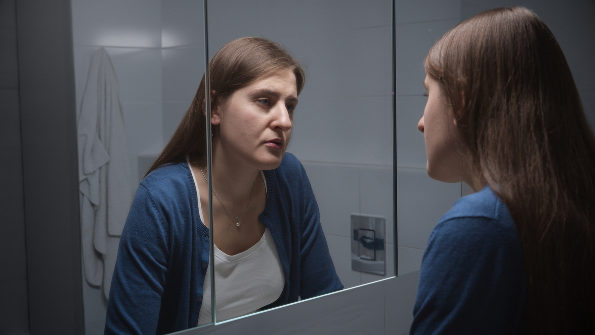 A stock image of an woman standing at sink  and looking at her reflection. 