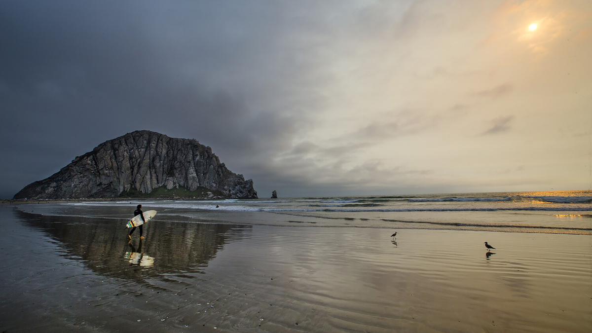 A surfer and Morro Rock are reflected in the water at low tide as the sun goes down with ash visible from a forest fire in Morro Bay in 2016. (Allen J. Schaben/Los Angeles Times/TNS)
