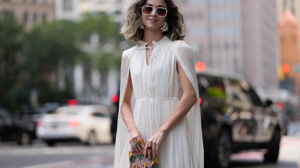NEW YORK, NEW YORK - SEPTEMBER 13: a guest wears shoulder-length wavy hair with darker roots and lighter ends, white-framed sunglasses with pink lenses, a small multicolored beaded clutch, a sleeveless ivory pleated maxi dress with a tie keyhole neckline and attached cape panels, a cinched waist and tiered ruffled hem, outside Altuzarra, during New York Fashion Week, on September 13 2025 in New York, New York (Photo by Edward Berthelot/Getty Images)