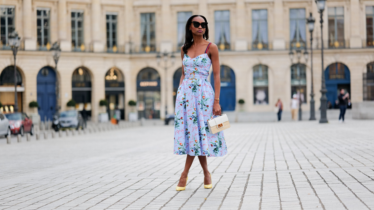 PARIS, FRANCE - APRIL 29: Emilie Joseph wears black large sunglasses, crystal earrings, a pale blue with red / green / navy blue / beige flower print pattern heart neck / tank-top / belted / midi dress, gold bracelets, a beige wickers and white shiny leather bi-material handbag, yellow shiny leather pointed pumps heels shoes , during a street style fashion photo session, on April 29, 2023 in Paris, France. (Photo by Edward Berthelot/Getty Images)