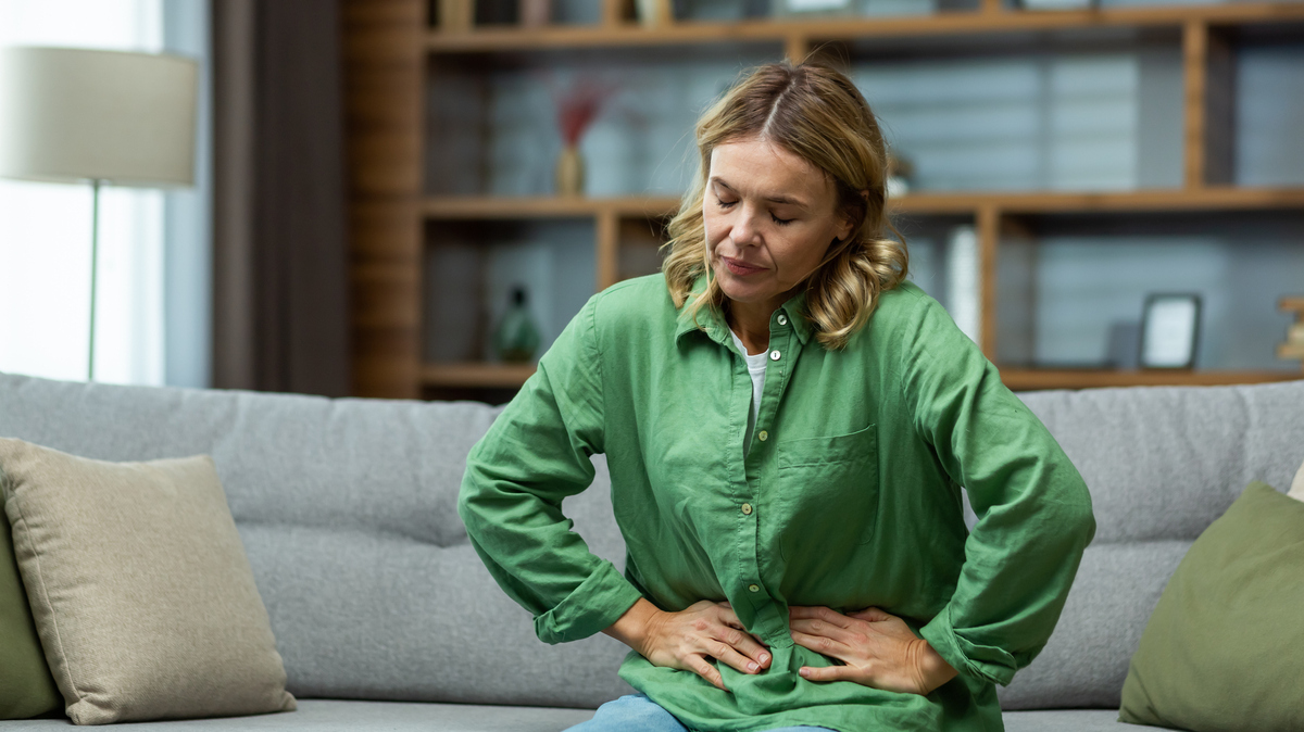 A stock image of a woman is sitting on the sofa at home, holding her stomach with her hands. 