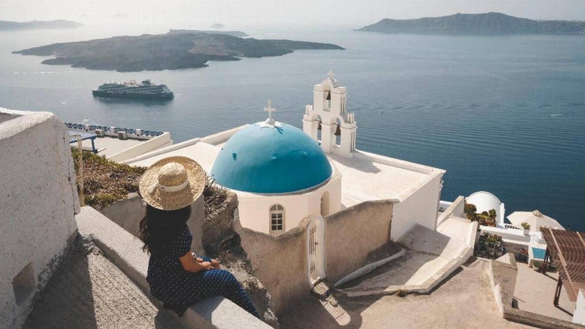 A woman sits on a ledge in Santorini with the Celebrity Apex cruise ship anchored in the distance. 