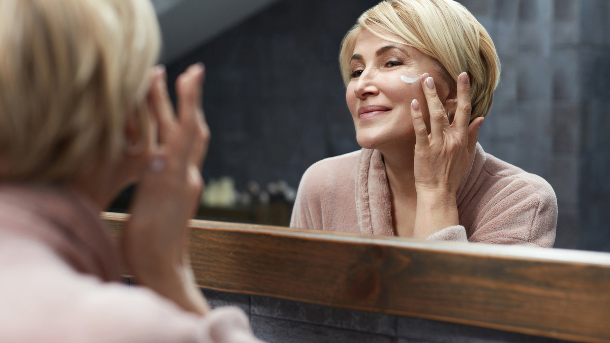Skincare Routine. Mature Woman Uses Cosmetic Cream On Face Skin In Front Of The Mirror Reflection. Anti Aging Beauty Treatment Concept.