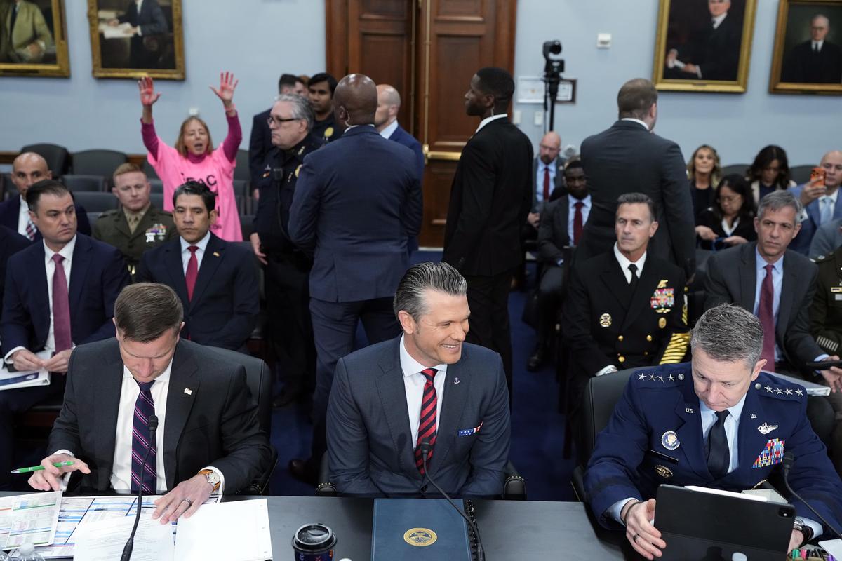 Secretary of Defense Pete Hegseth smiles as security officials prepare to remove Medea Benjamin, founder of the anti-war group Code Pink, from the room prior to the start of a hearing before the House and Senate appropriations subcommittees on defense in Washington on Tuesday, May 12, 2026. Hegseth is flanked by Undersecretary of Defense Jules Hurst, left, and Gen. Dan Caine, the chairman of the Joint Chiefs of Staff. (Haiyun Jiang/The New York Times)