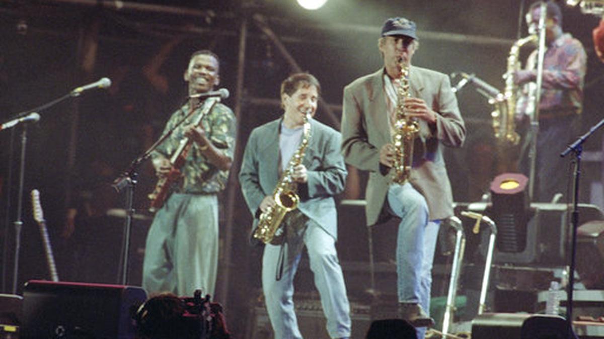 FILE - In this Aug. 16, 1991 file photo, Paul Simon, center, plays a finale with lead guitarist Ray Phiri, left, and actor-comedian Chevy Chase on the saxophone in New York’s Central Park. Ray Phiri, a South African jazz musician who founded the band Stimela and performed on Paul Simon’s Graceland tour, died of cancer on Wednesday July 12, 2017 at the age of 70.