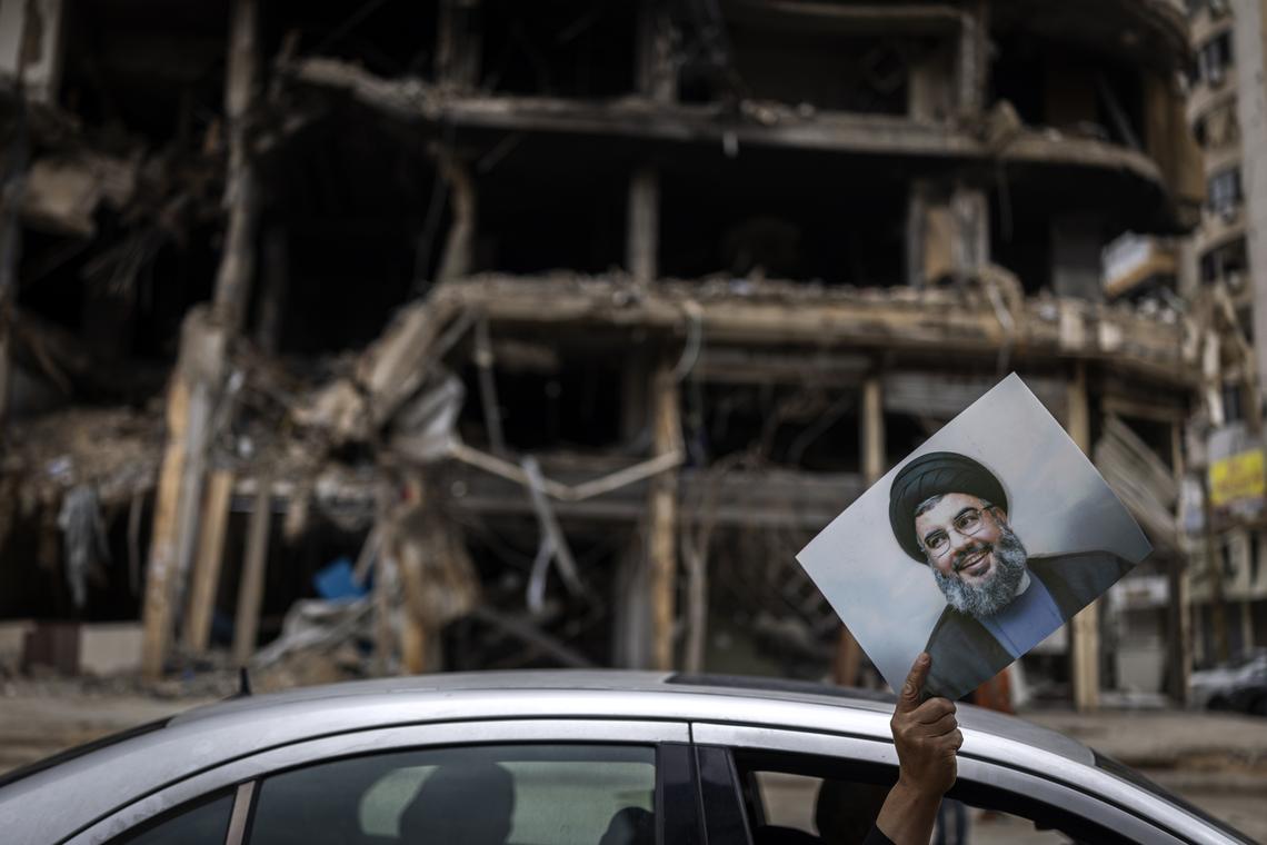 A man holds up a portrait of the slain Hezbollah leader Hassan Nasrallah as he is driven past the wreckage of a building destroyed in an Israeli airstrike in Dahiyeh, the suburb south of Beirut, Lebanon, on Monday, April 20, 2026. Joseph Aoun, the Lebanese president, said that he had appointed Simon Karam, a former ambassador to the United States, to lead talks with Israel aimed at ending the war and achieving a complete Israeli withdrawal from southern Lebanon. (Diego Ibarra Sánchez/The New York Times)