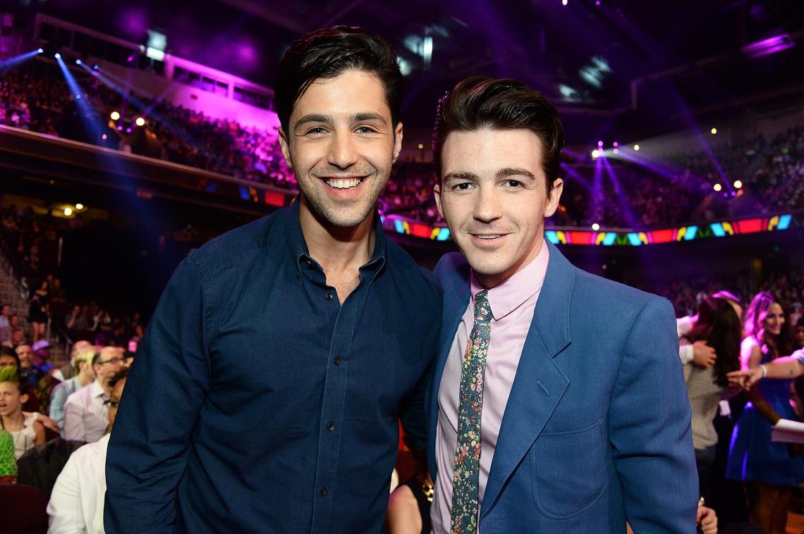 A man with a wide smile and a dark blue shirt, Josh Peck, stands next to another man with a pink shirt and a blue blazer, Drake Bell. Both men are looking at the camera and are posing in what appears to be a large indoor arena or stadium with a crowd in the background.