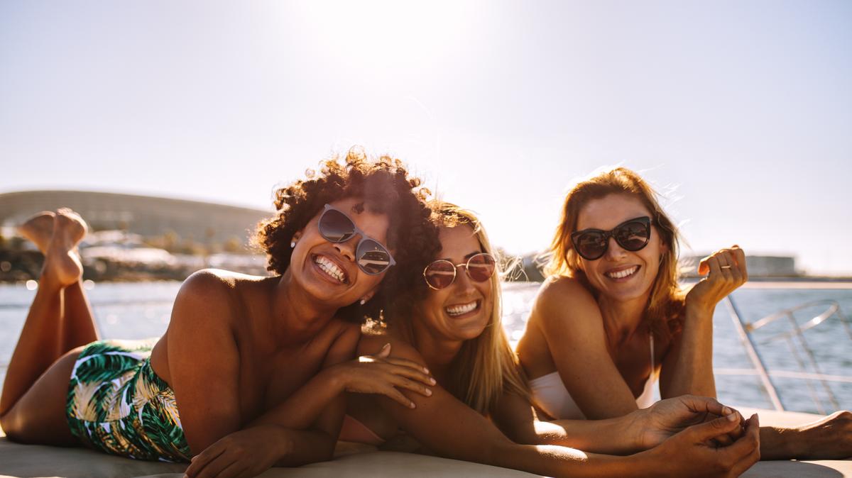 Group of beautiful women relaxing on a yacht deck. Three female friends sunbathing on small boat looking at camera and smiling.
