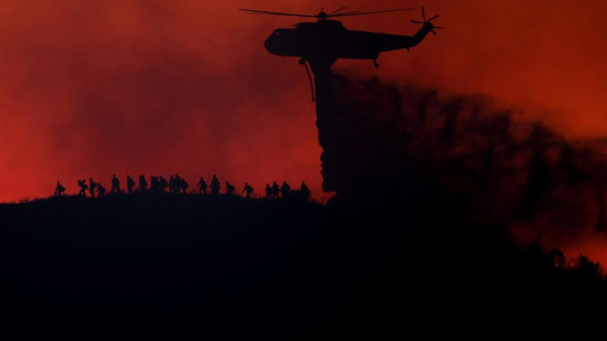 A helicopter drops water as firefighters walk along a ridge during the Sandy fire in Runkle Canyon on May 19, 2026, in Simi Valley, California. (Eric Thayer/Los Angeles Times/TNS)
