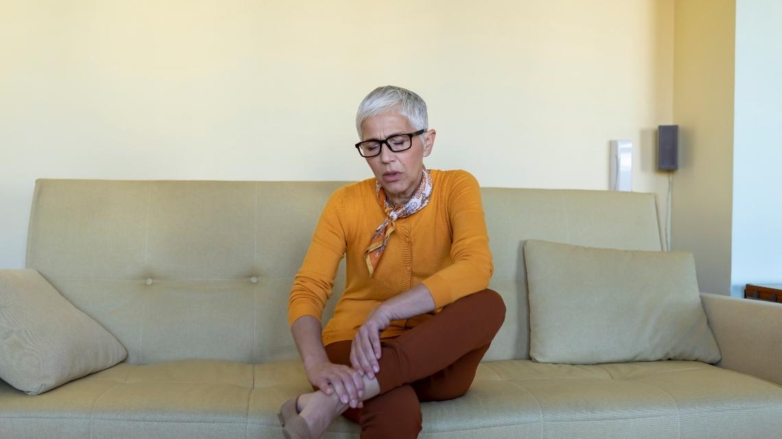 An Elderly Woman is Sitting on the Sofa at Home and Holding her Leg Wrist due to Insufferable Pain. Photo of an Elderly Woman Feeling Hard Pain in her Leg.