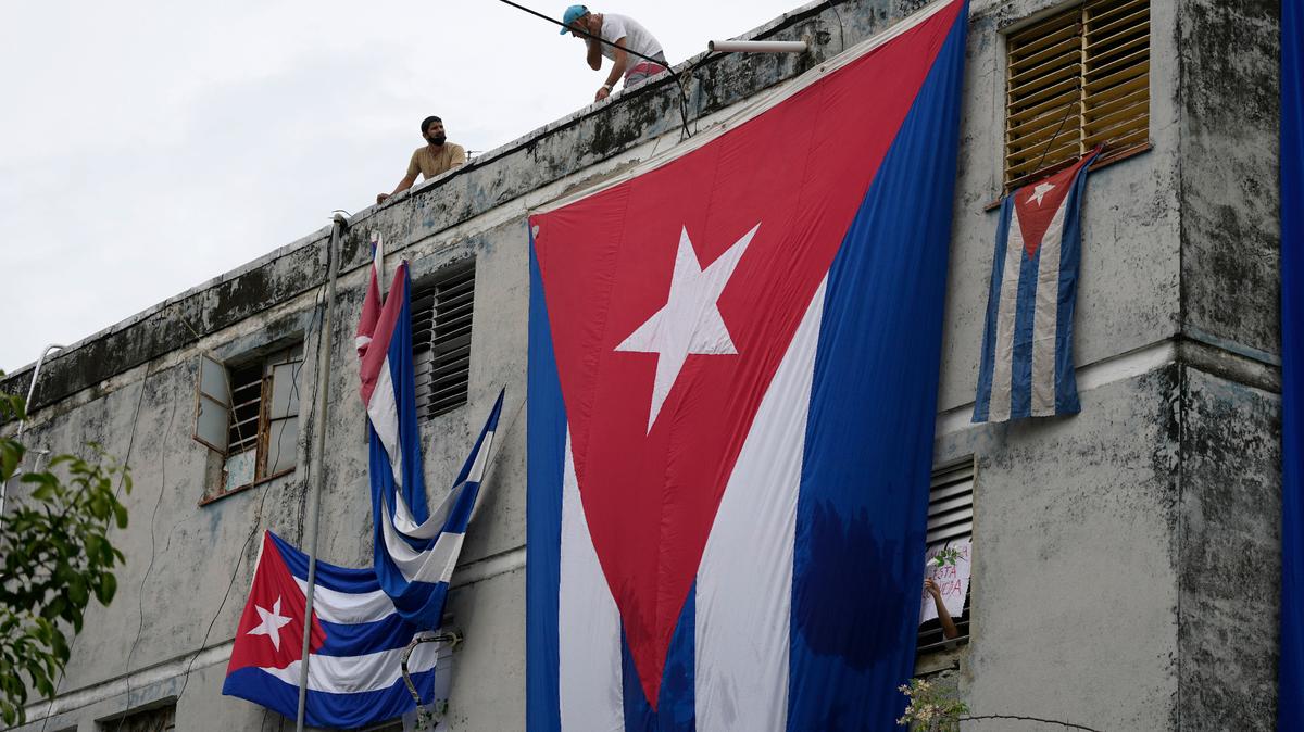 Hombres cuelgan banderas cubanas en las ventanas de la casa del dramaturgo y opositor Yunior García en un intento de evitar que se comunique con el exterior, mientras sostiene una flor desde una ventana, en La Habana, Cuba, el domingo 14 de noviembre de 2021. (AP Foto/ Ramón Epinosa)