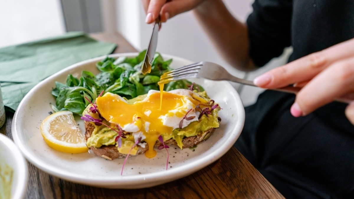 Young woman eat breakfast with knife and fork, Morning in cafe. Healthy breakfast with wholemeal bread toast with avocado, poached egg with green salad.
