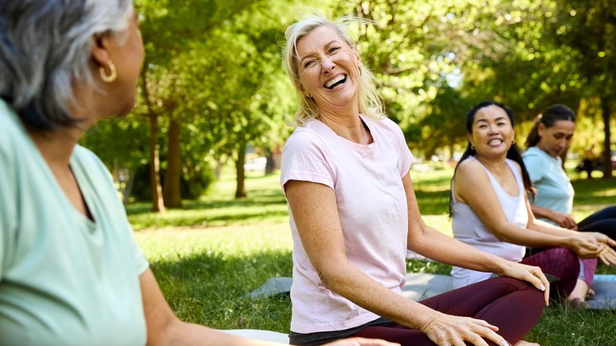 Happy woman sitting for yoga with friends. Mature female is laughing while enjoying morning routine. She is in public park.