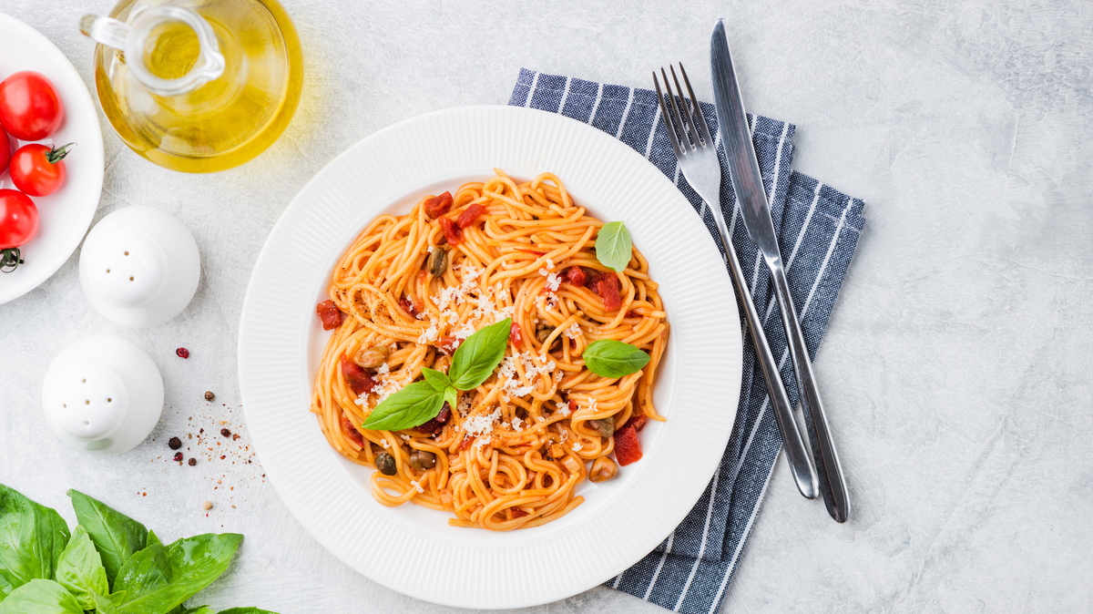 Italian pasta spaghetti with sun dried tomatoes, basil and cheese on white plate, grey concrete backdrop, top view