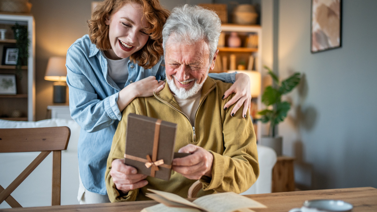 Loving granddaughter is embracing her grandfather and giving him a gift while he's sitting at the table, reading a book, in a cozy home interior