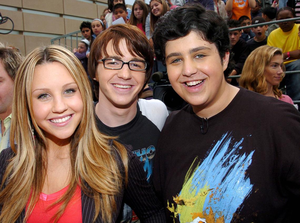 A smiling group of three young actors: Amanda Bynes on the left with long blonde hair, Drake Bell in the middle with glasses and brown hair, and Josh Peck on the right with dark, curly hair. They are standing close together and appear to be posing for a photo in a public space, with a crowd of people visible in the background