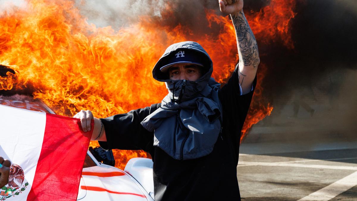 A protester raises a fist while holding a Mexican flag in front of a Waymo vehicle that was set on fire during a demonstration following federal immigration operations in Los Angeles on June 8, 2025.
