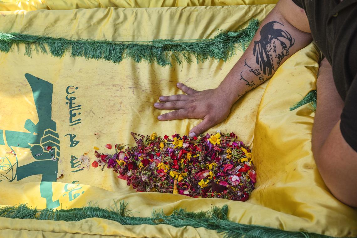 A grieving man, with a tattoo of the late Hezbollah leader Hassan Nasrallah on his arm, touches a coffin draped in the Hezbollah flag during a mass funeral for 20 people in the southern Lebanese village of Qlaileh, on Tuesday, April 21, 2026. The cease fire has given families who had fled because of Israeli evacuation warnings the opportunity to return to the village and bury their relatives killed during the war after being temporarily buried in transit cemeteries. (David Guttenfelder/The New York Times)