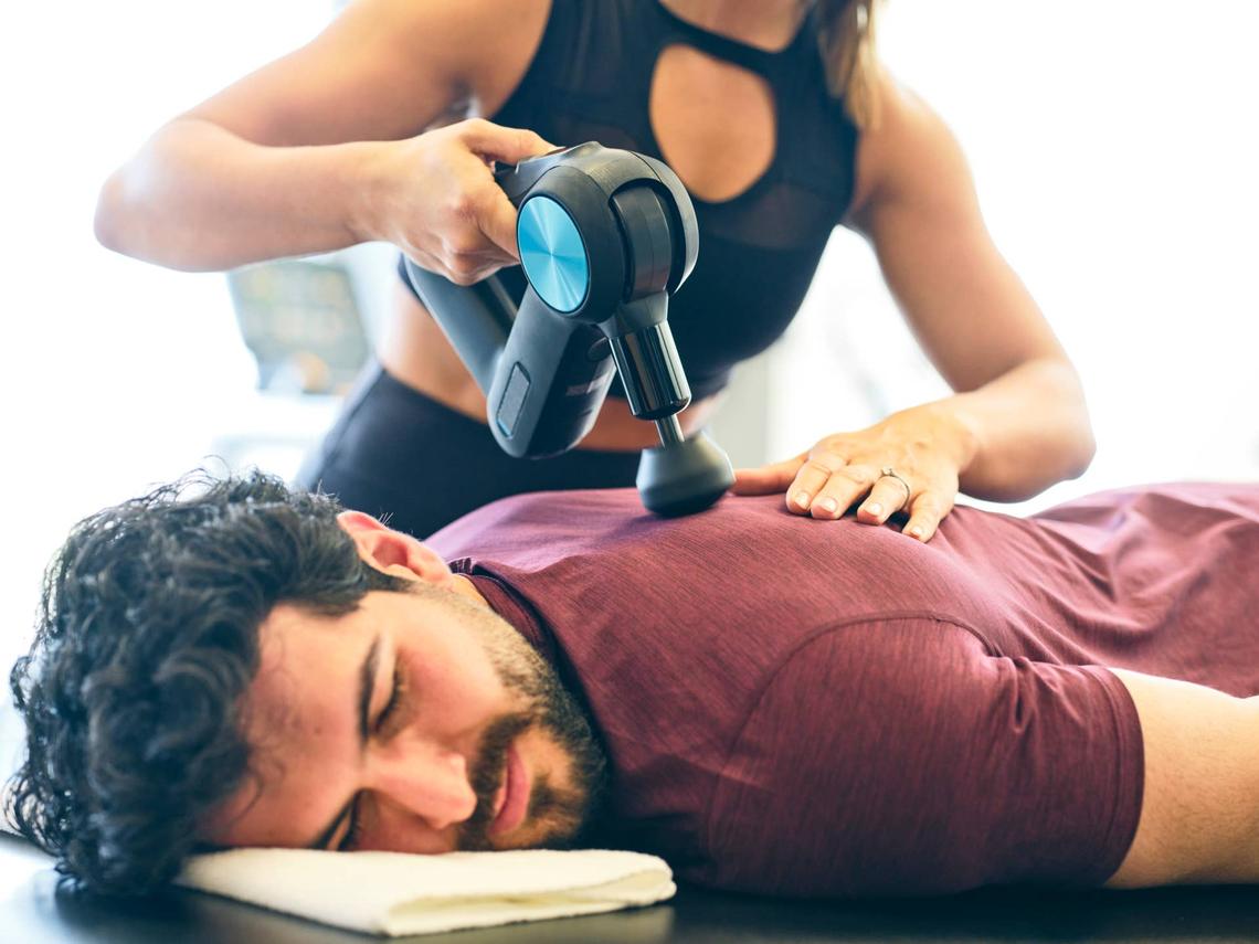  A fitness trainer massaging an athlete with a massage gun in a gym. 