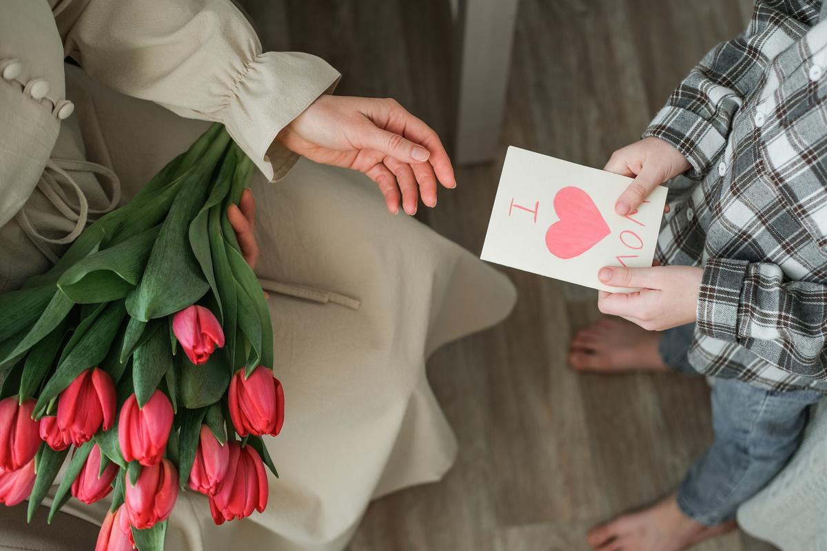  Child boy giving a handmade “I Love Mom” card to mother holding red tulips. Top view of heartwarming family moment, perfect for Mother’s Day, birthday or spring holiday concepts 