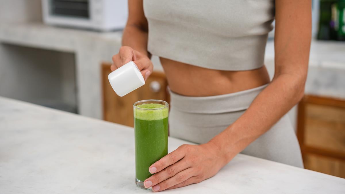 An unrecognizable woman is adding a protein powder in a glass of green smoothie.