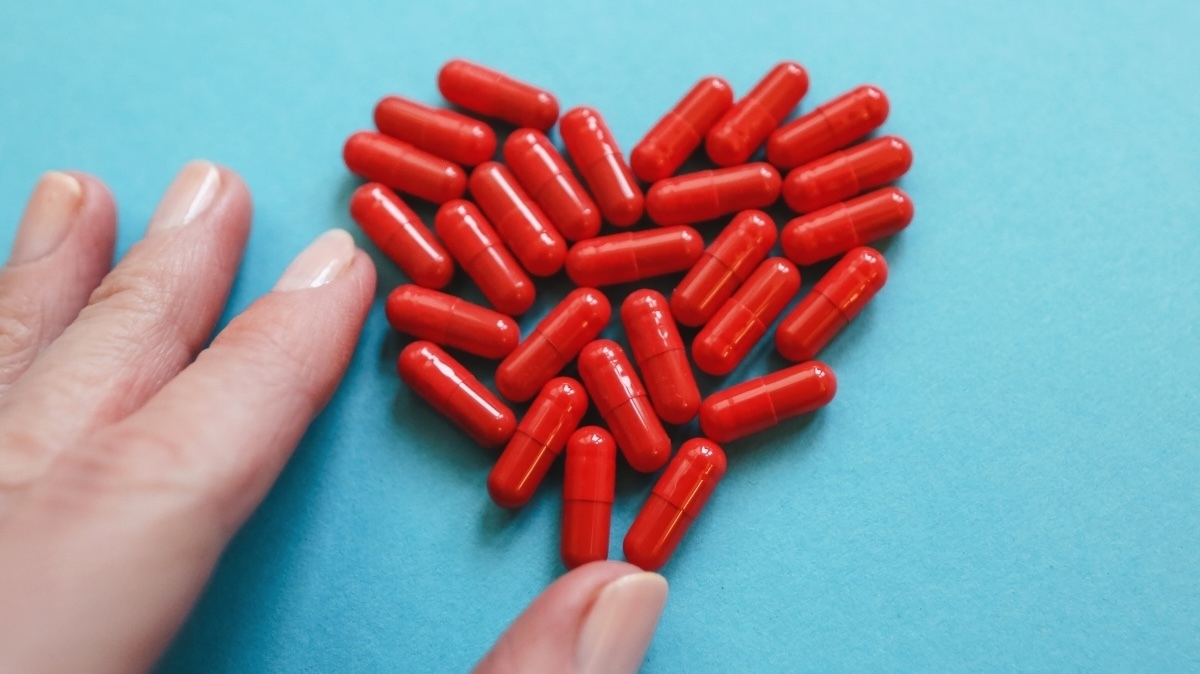 Healthcare And Medicine. Cropped Woman's Hand Holding Small Red Iron Capsule For Support And Prevention Anemia Against Heart Shaped Heap Of Pills Background