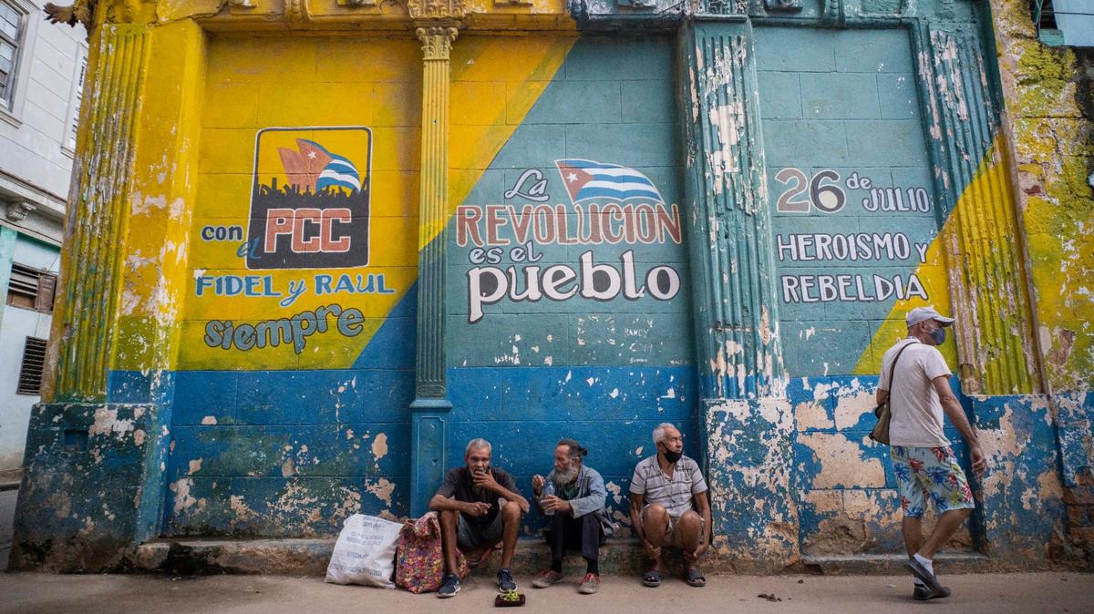 ARCHIVO - En esta fotografía de archivo del 23 de diciembre de 2020, personas con máscaras mal ajustadas utilizadas contra la propagación del nuevo coronavirus se sientan en una acera junto a un muro con consignas políticas en La Habana, Cuba. (AP Foto/Ramón Espinosa, Archivo)