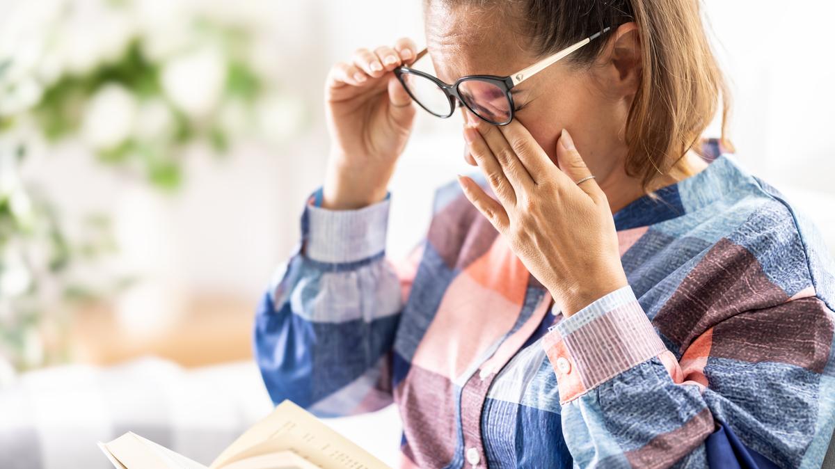 A woman sits on a sofa with an open book and tiredly massages the bridge of her nose under her glasses with both hands. She has a headache or severe eye fatigue while reading.