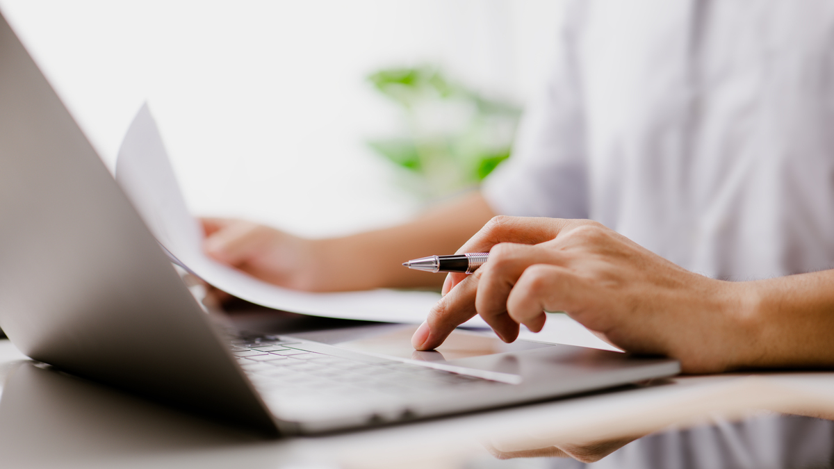 A stock image shows a   hand typing on a laptop keyboard, while reading a business document. 